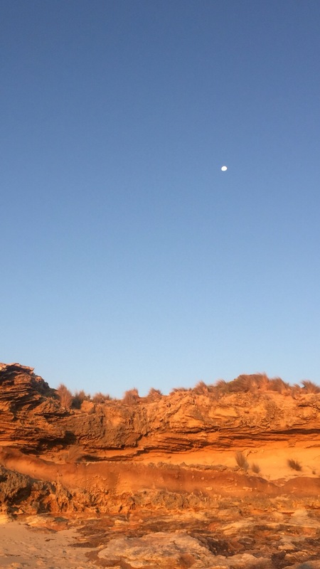 This is a picture of the moon in the sky during the sunset at a beach. 