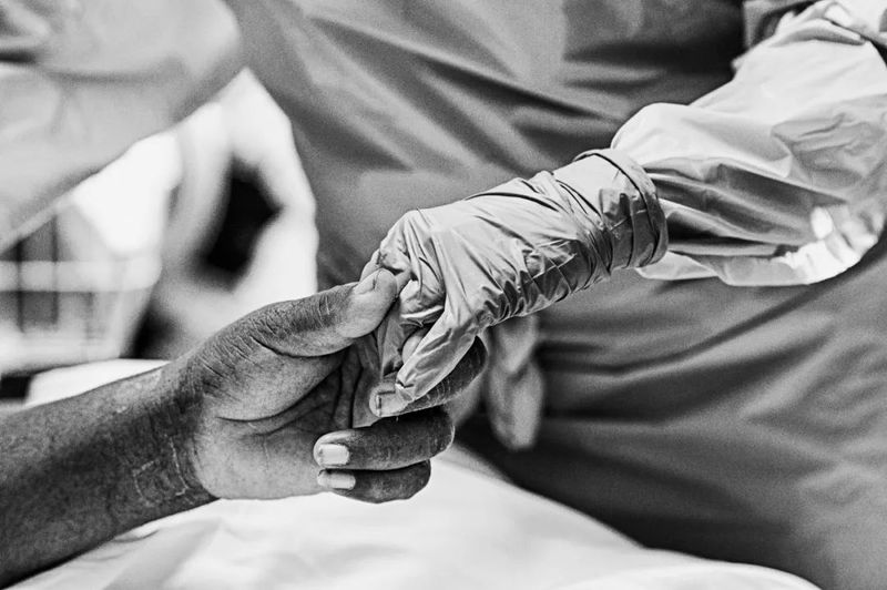 Black-white image of a dark-skinned hand holding the gloved hand of a person who is in full surgical gear. 