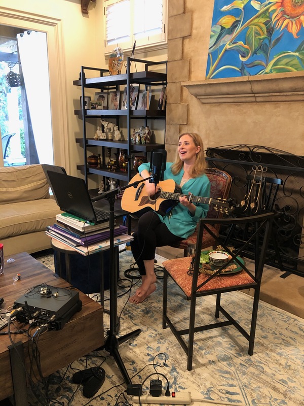 A woman in a residential setting playing the guitar and singing near a microphone while a computer faces her.