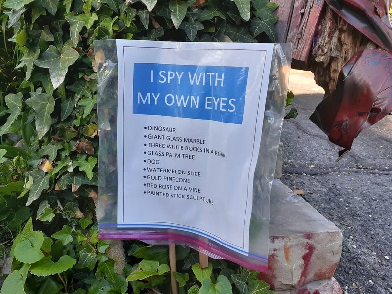 This is a picture taken of a sign in a yard which reads "I spy with my own eyes: dinosaur, giant glass marble, three white rocks in a row, glass palm tree, dog, watermelon slice, gold pinecone, red rose on a vine, painted stick sculpture."