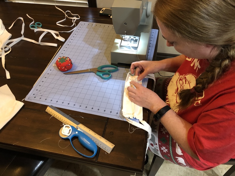 A person wearing a red shirt is sewing a white face mask with a green sewing machine on top of a wooden table. There are white scraps all over the table. 