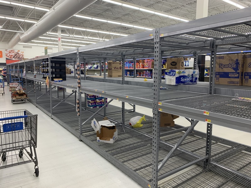 Empty rows of grey shelves in an isle. Looking through the shelves there are a few rolls of paper towels. 