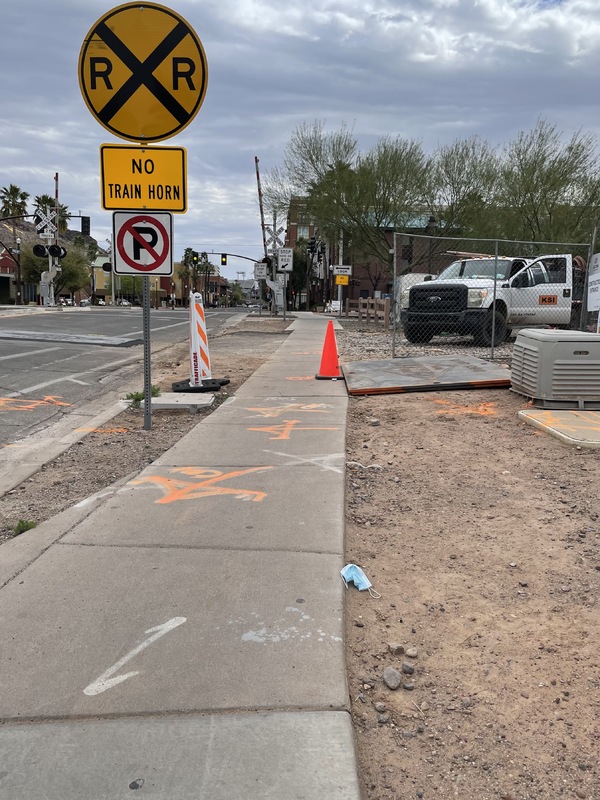 This is a picture of a discarded face mask that has been left near a sidewalk. A railroad crossing, several cars, and some buildings can been seen in the background. 