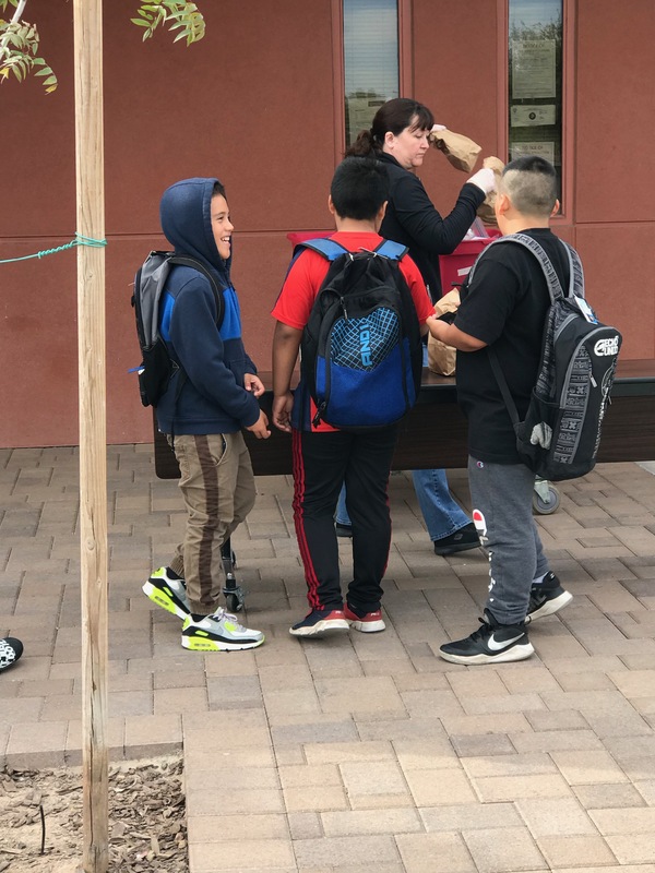 Three kids wearing backpacks are standing in front of a table outside. The kids are talking to one another. Behind the table is a woman who is putting paper lunch bags onto the table. 