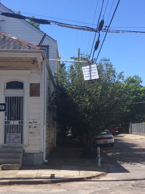 Corner of a street with an older house with a sign in the door with text, "BALANCE? Where's The Fairness?" with a photo of a scale and it tipping to one side.  Another sign hanging from a pole with text, "How Many Thousands More Americans Will Die....Due to Trump's Horrific Arrogance?"