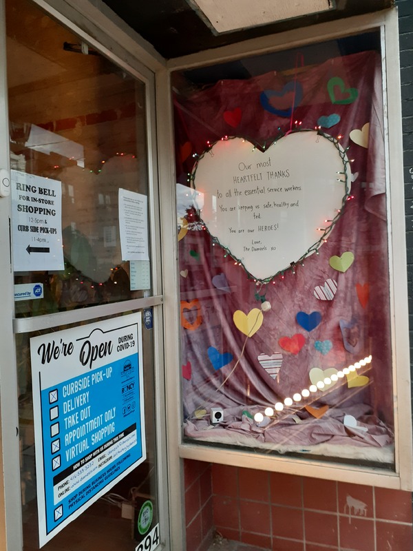 This is a picture taken of a storefront window, where several signs are present. One sign lists curbside pickup, appointment only, and virtual shopping as the only options to purchase items from the store. A paper heart in the window reads: "Our most heartfelt thanks to all the essential service workers. You are keeping us safe, healthy, and fed. You are our heroes! Love the Daniels xoxo."