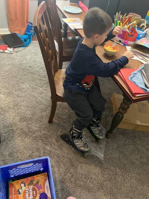 This is a picture of a young boy seated at a table and eating some food. There are papers and pencils covering the rest of the table.