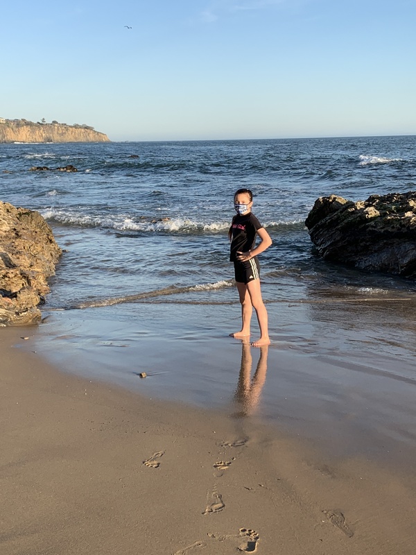 This is a picture of a young girl wearing a face mask with her back to the ocean at the beach. 