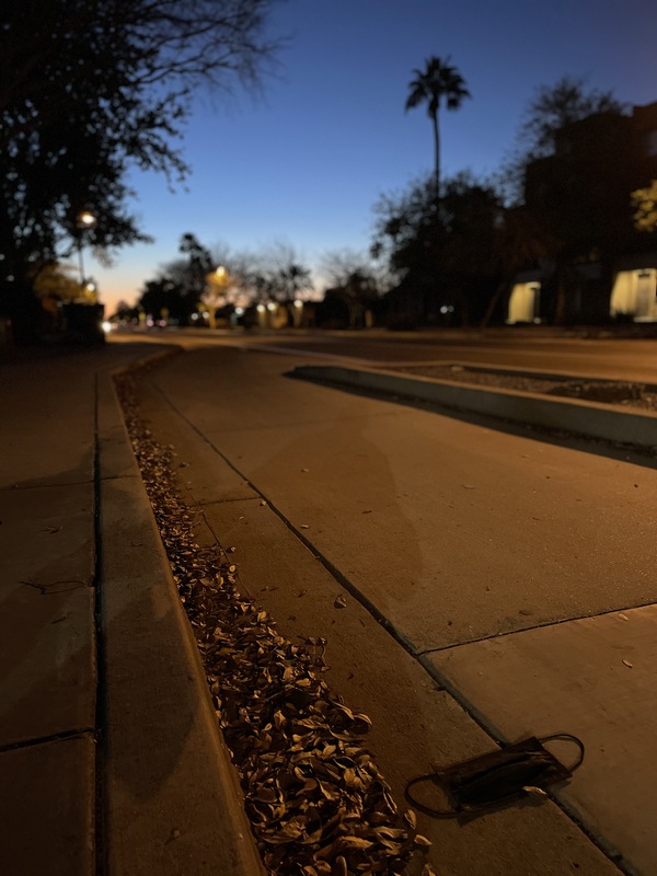 This is a picture taken of a discarded cloth face mask resting on a city street as the sun sets in the background. 