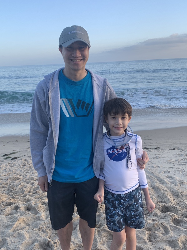 This is a photo taken of a young boy and a man standing side by side at the beach, with the ocean behind them. 