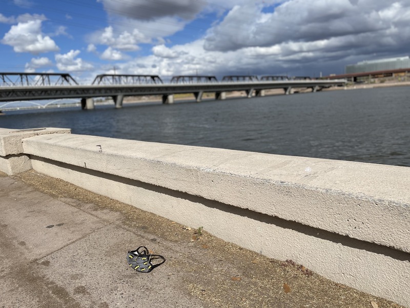 This is a picture of a batman themed face mask that has been discarded in the dirt at the edge of a lake. A rail bridge can be seen in the background. 