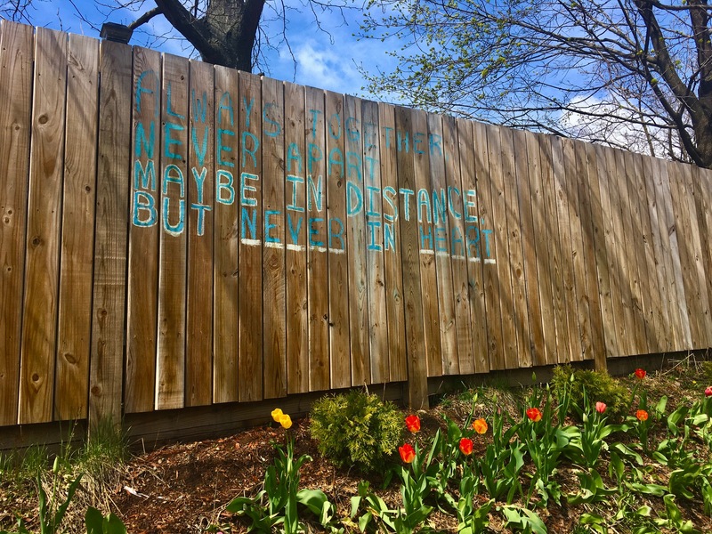 A message painted on a fence meant to brighten the day of the reader that says "Always Together, Never Apart, Maybe In Distance, But Never In Heart". 