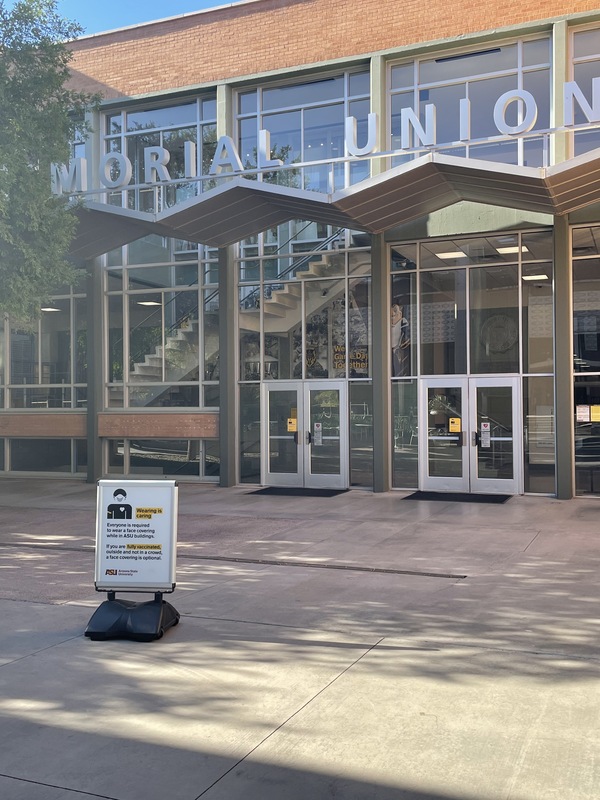 This is a picture taken outside the ASU Memorial Union. A sign that has been put up in front reading: "Everyone is required to wear a face covering while in ASU buildings. If you are fully vaccinated, outside, and not in a crowd, a face covering is optional." 