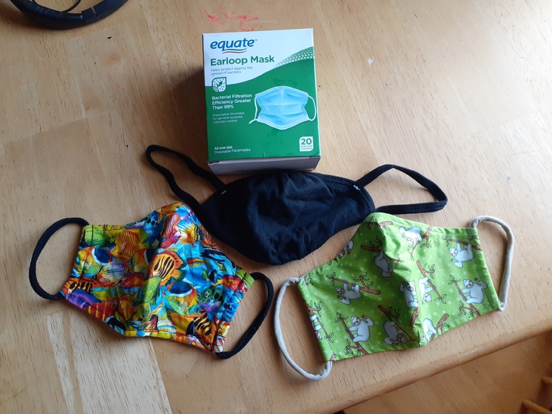 This is a picture of three different cloth face masks sitting on a table, with a box of twenty disposable masks sitting next to them. 