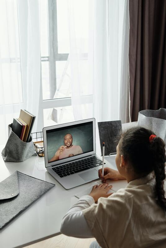 This is a picture of a girl writing in a notebook while she watches a person talk on a video pulled up on her laptop screen. 