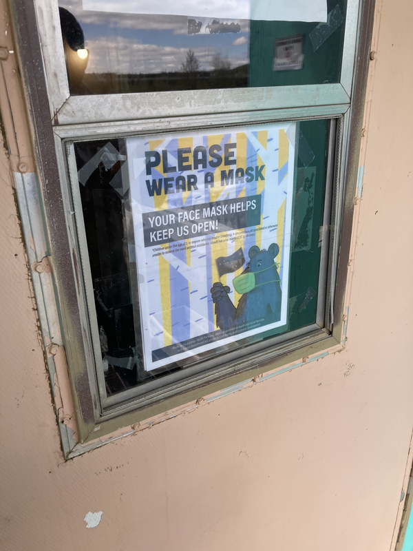 This is a picture of a sign which depicts a bear wearing a face mask and waving a flag. The sign reads: "Please wear a mask. Your face mask helps keep us open!"