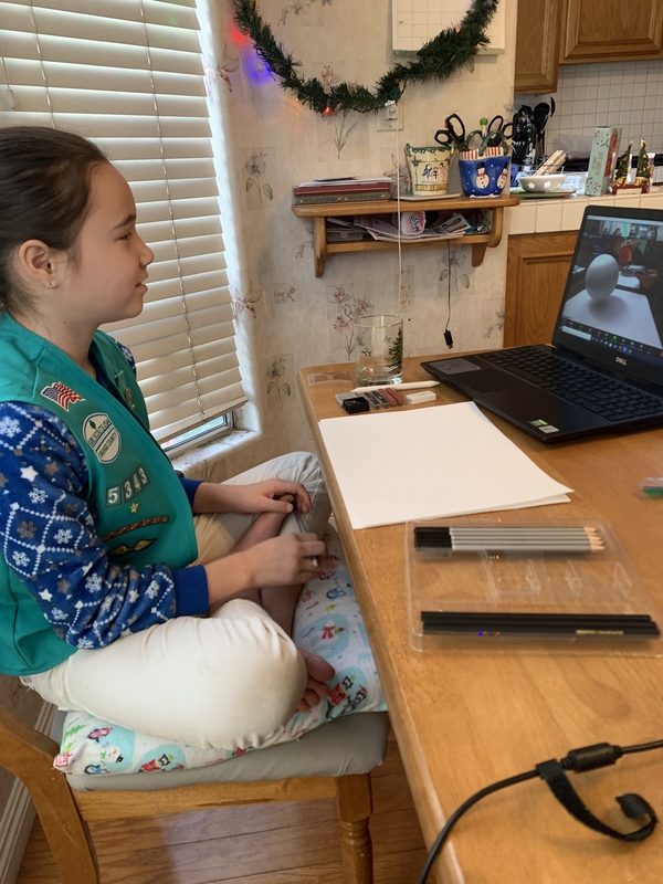 Young girl with Girl Scout vest in front of computer with paper and drawing materials.