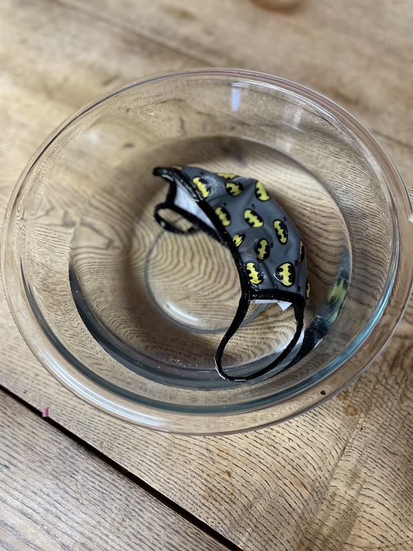 This is a picture of a batman themed face mask soaking in a glass bowl of water on a table. 