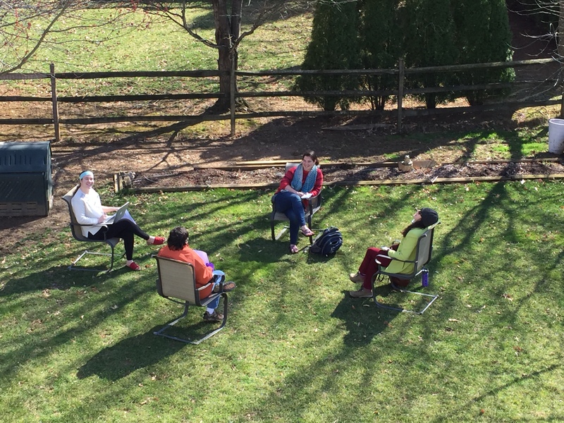 Four people that are 6 feet apart are sitting in outdoor chairs. The person on the far left has a laptop in their lap. 