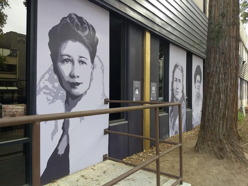 This is a picture taken of a series of murals that are painted on the side of a building. Various women in different periods of clothing are depicted in black and white. 