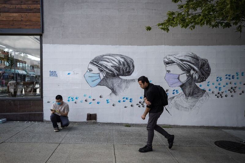 One man walking past a mural of two women wearing masks surrounded by flying birds. Another man crouches next to the mural on his phone. 