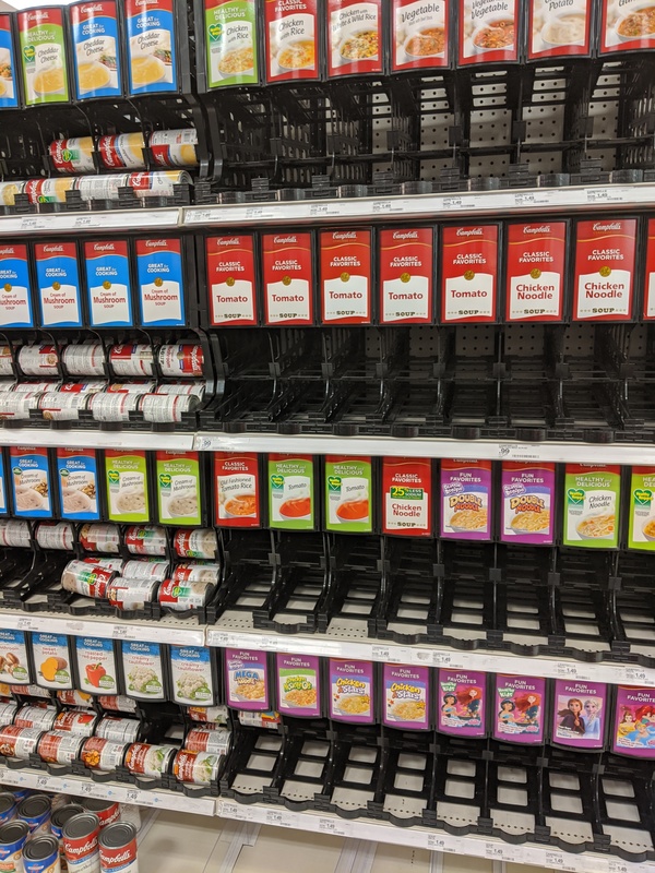 A row of numerous shelves that hold cans of soup. On the right of the picture has empty shelves that used to have tomato soup, chicken noodle, vegetable soup, potato soup, and chicken and rice soup. On the right of the picture shows shelves stocked full of soups in a can which are cheddar cheese soup, cream of mushroom, and various cans of vegetables on the bottom shelf like sweet potato, creamy cauliflower, and roasted red pepper. 