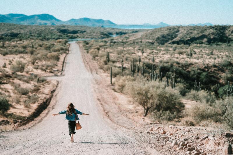 This is a picture of a woman in a dress, carrying a handbag while walking down a dirt road in the desert with her arms spread wide. 