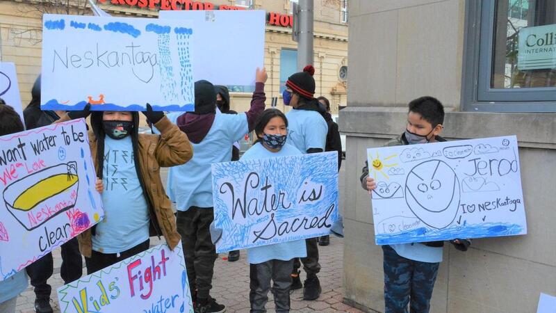 This is a picture of face masked children holding signs that protest the Neskantaga water crisis. One sign reads "water is sacred." 