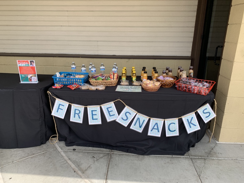 This is a picture of a table, which has a sign on it reading "free snacks". Various foods and drinks to consume are set out on the table. 