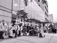 This is a picture of a group of doctors kneeling on the ground in solidarity with the Black Lives Matter movement. A large glass building is being them. 