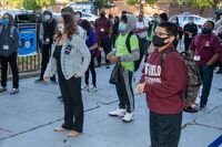 This is a picture taken of a group of people waiting in line on the street while social distancing. Every person is wearing a face mask.