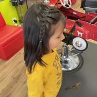 This is a picture taken of a girl that has just had a haircut. Her shoulder length hair is partially braided, and she is wearing a yellow shirt with blue pants. 