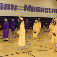 Image of soon to be high school graduates in their caps and gowns, standing spread out in a gym.