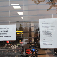 Two signs at shop.
The first sign says: "We love you but... No hugging, no kissing, no handshaking."
The second sign says: "Closed. We will reopen as soon as possible." and then gives the shop's website link along with associate's e-mail addresses and work phone numbers. 