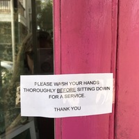 A lamented rectangle sign taped on a bright pink door that says: PLEASE WASH YOUR HANDS THOROUGHLY BEFORE SITTING DOWN FOR A SERVICE. THANK YOU.  