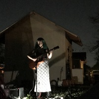 This is a picture taken of a woman who is performing music on a stage outdoors at night. She is wearing a white dress and holding a guitar, with a spotlight on her that silhouettes her picture against the wall behind her.  