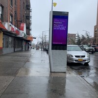 this is a picture of a 'LinkNYC" electronic advertisement kiosk on the street, which has a message on it reading: "COVID-19 Info, Prevention. Stay home if possible, even if you are healthy."