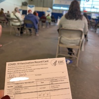 This is a picture taken of a person's vaccine card, with other people waiting to leave the vaccination area in the background. 