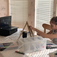 Child sits at a table in front of an open laptop screen of a Zoom art class with a tabletop easel and canvas.