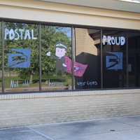 Window Writing Reading "Postal Proud. Not all Heroes Wear Capes" along with a drawing of a postal service man.