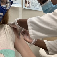 This is a picture of a woman wearing a face mask receiving her first COVID-19 vaccine shot from a healthcare worker. 