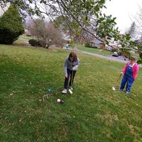 Two women are photographed on a large lawn playing a game of croquet. One is reaching for a ball, while another watches from the background.
