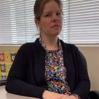 This is a picture of a women wearing a black coat staring quizzically at the camera as she sits at a table.