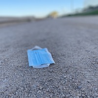 This is a picture of a blue face mask that has been discarded on a gravel and dirt path. The rest of the background is out of focus. 