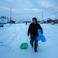 This is a picture of a man in dark winter clothing dragging a sled in the snow behind him, and carrying three jugs. 