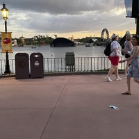 This is a picture of a face mask that has been discarded on a concrete walkway near a pier. Groups of people walk by in the background while the sun sets. 