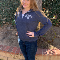 This is a picture of a girl wearing a UC Irvine sweatshirt and jeans smiling in front of a planting area with trees in the background. A UC Irvine logo is present in the left hand corner of the photo. 