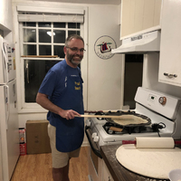 This is a picture of a man smiling while he cooks dough in a skillet in a kitchen. 