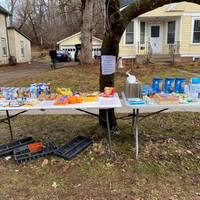 Two foldable white tables are laid out standing next to each in front of a yellow house. Behind the tables is a tree with a piece of paper nailed to it. On top of the foldable tables is various food items and cans. 