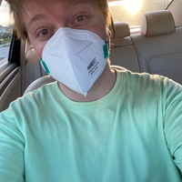 A selfie of a man wearing a green shirt and a surgical mask sitting in his car.